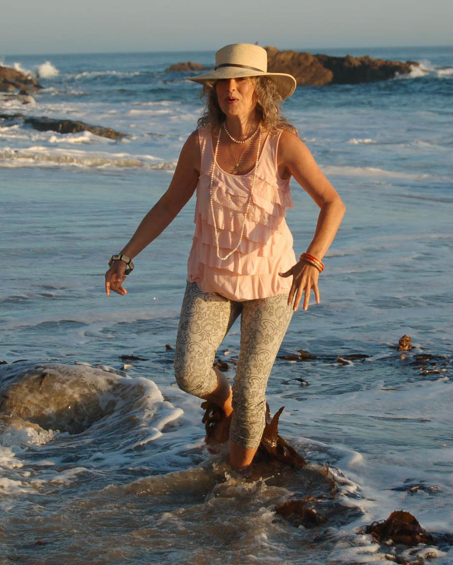Mischa Martineau walking in the ocean waves with a sun hat, peach colored top, leopard tights and a string of white pearls with ocean rocks in the background.