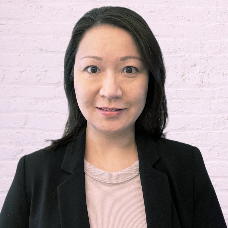 Christine Law professional headshot wearing a black business jacket, tan blouse on a white brick background