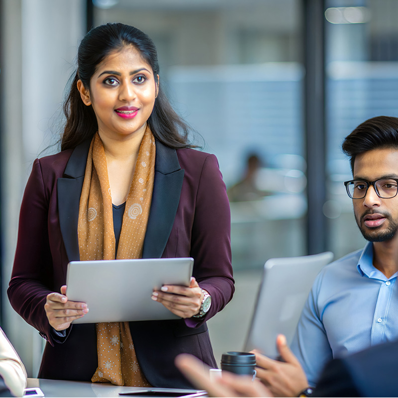 Indian woman leading a tech team presenting data from an ipad in a conference room with male colleagues